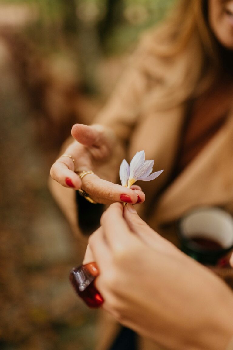 A close-up view of a woman holding a small flower, showcasing red manicured nails and a ring.