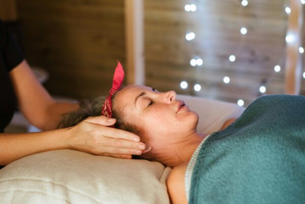 Side view of relaxed female patient with closed eyes lying on table under blanket while getting massage reiki during healing session in salon