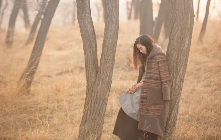 A girl draped in a warm coat deep in thought leaning against a tree in autumn