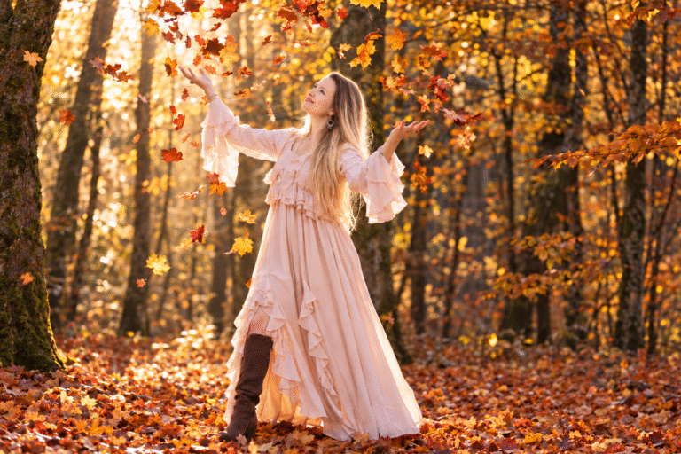 Corinne in a flowing blush-pink dress throwing autumn leaves in a sunlit golden forest